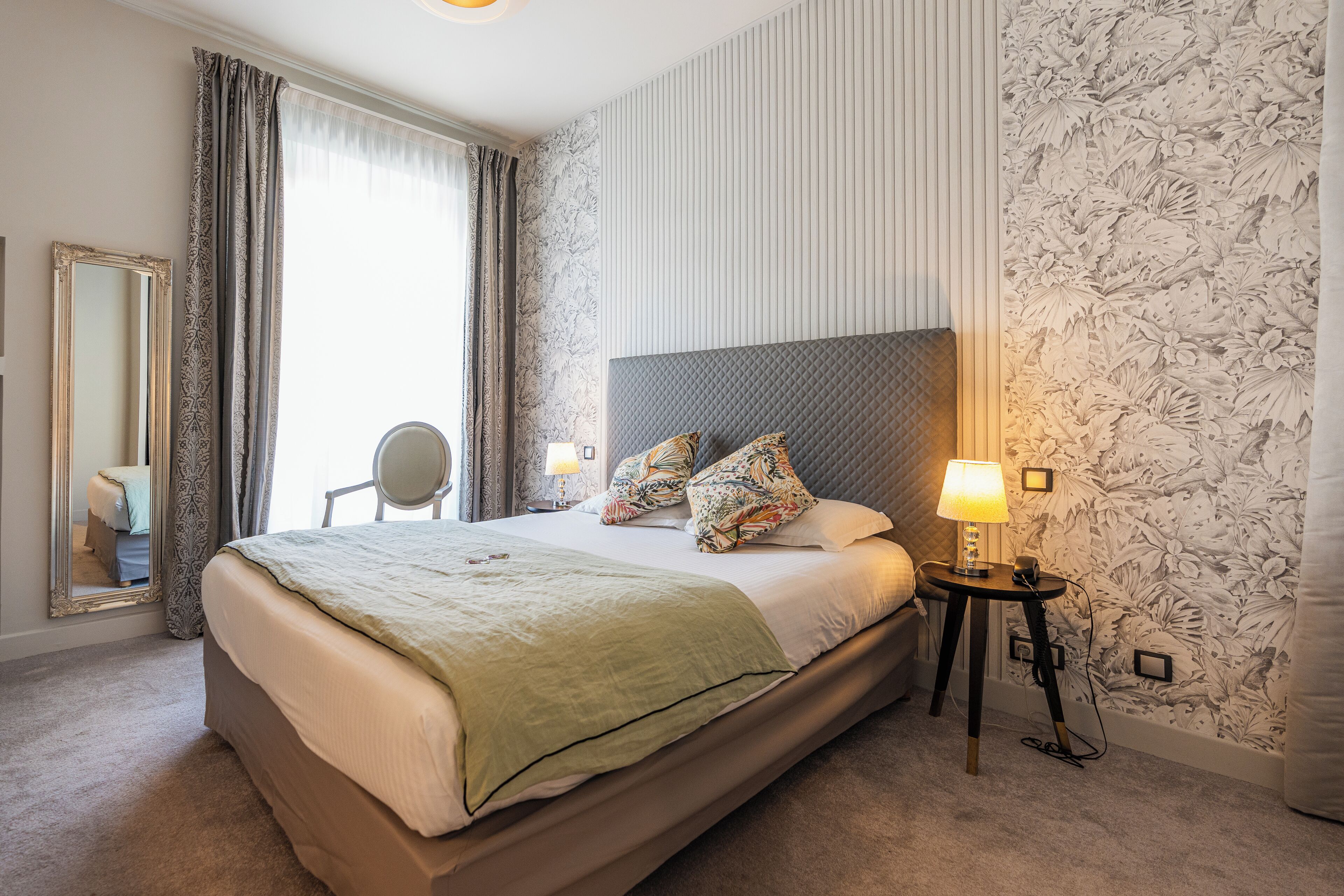 Een keurig opgemaakt hotelkamer met een groot bed, een nachtkastje met een lamp, een spiegel en bloemenbehang.