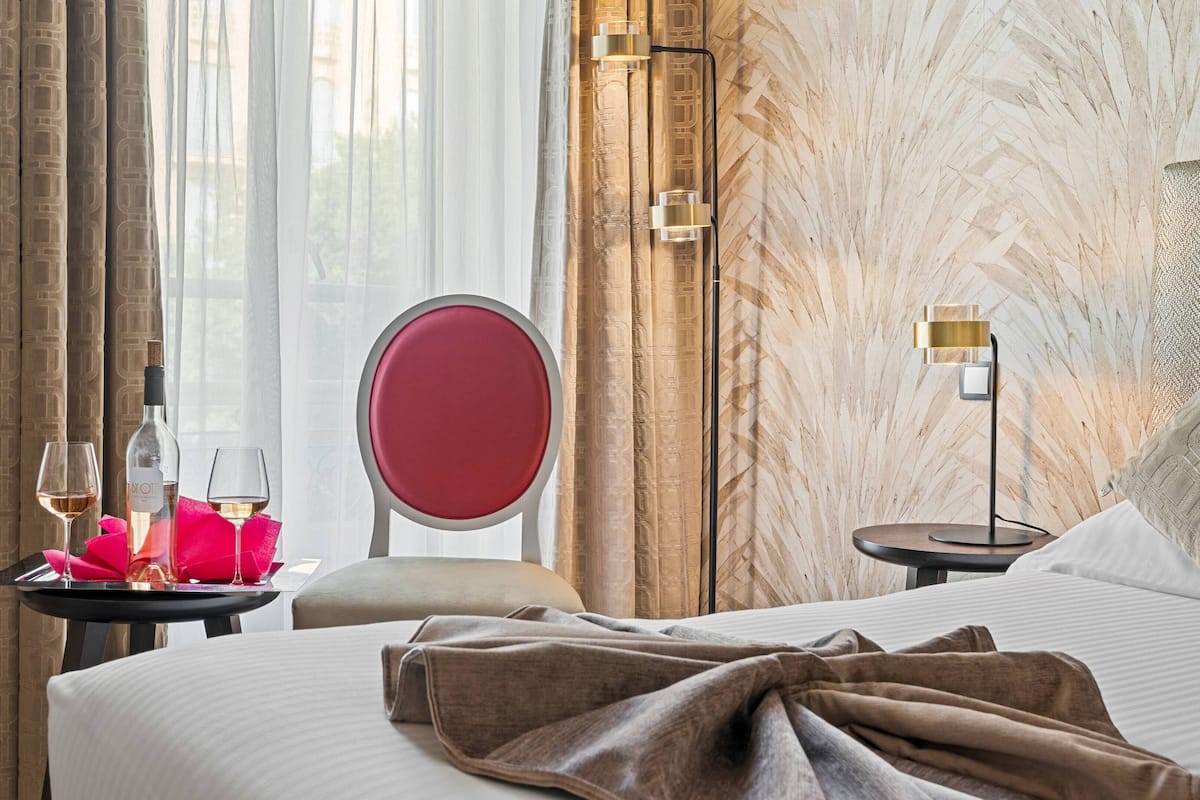 A hotel room with a bed, a bedside table with a lamp, a chair, a mirror, and a window with curtains.