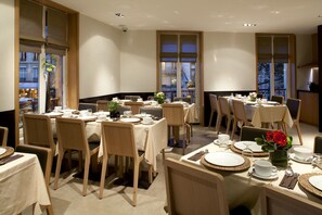 Un espace repas avec plusieurs tables dressées pour un repas, présentant un aménagement intérieur moderne avec des chaises et des tables en bois, et de grandes fenêtres offrant une vue sur le paysage urbain.
