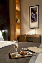A hotel room with a bed, a desk with a computer, a framed picture on the wall, and a tray with a glass of wine and cookies.