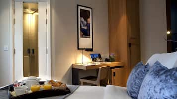 A hotel room with a bed, a desk with a laptop, a tray with breakfast items, and a framed photo on the wall.