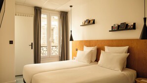 Una habitació d'hotel amb dos llits, un panell de capçalera de fusta, una petita prestatgeria amb llibres i una vista per una porta amb cortines.