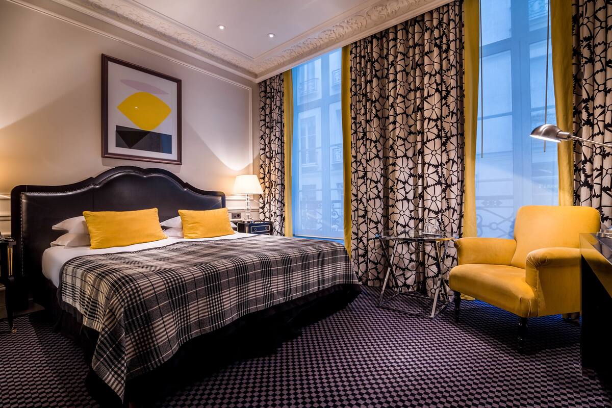 A hotel room with a large bed, a chair, a desk, and a view of the city through large windows.