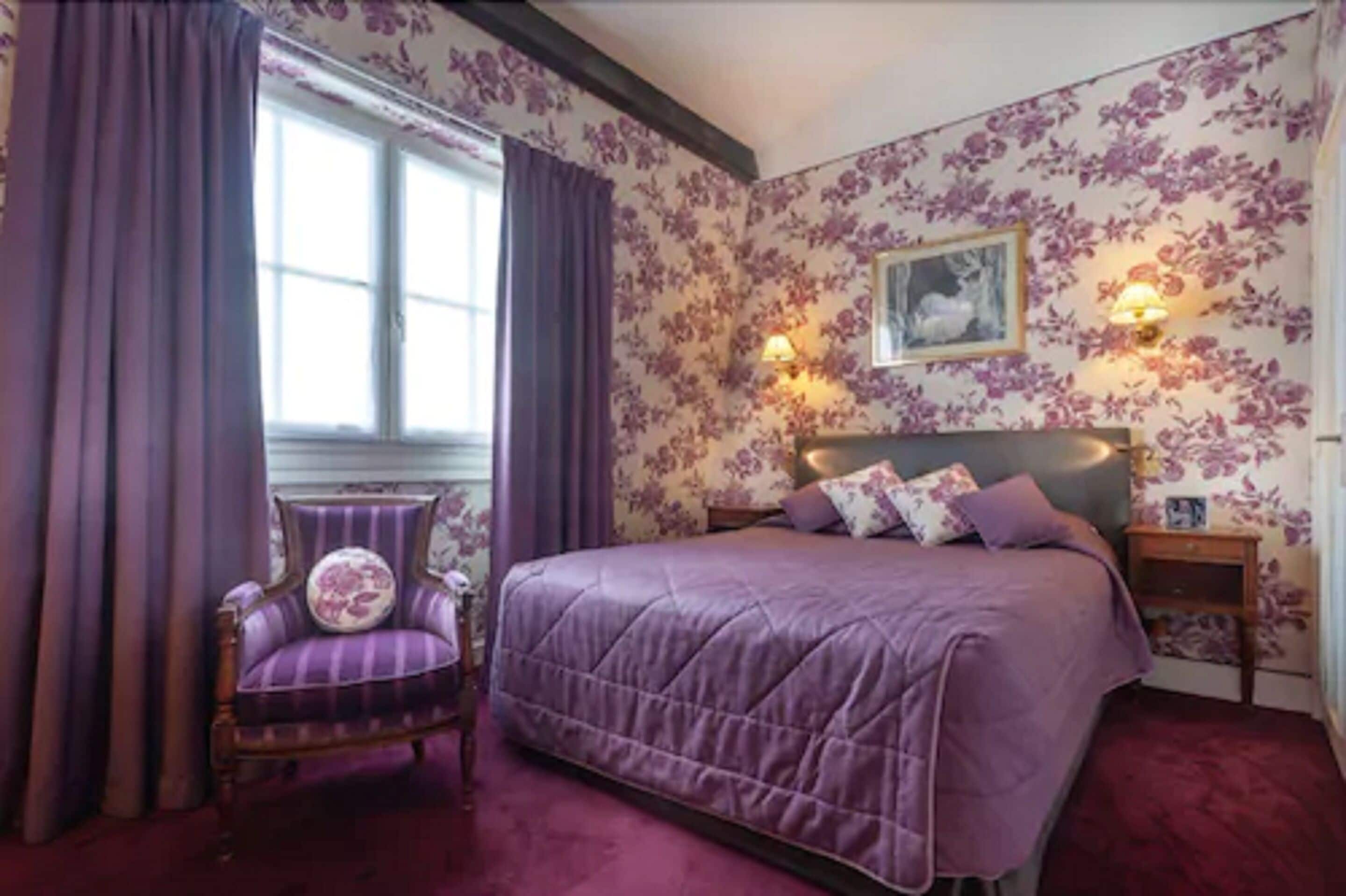 A bedroom with a large bed, purple bedding, a chair, and floral wallpaper.