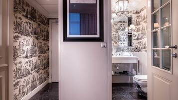A hallway with black and white patterned wallpaper, a framed picture on the wall, and a bathroom with a sink, mirror, and toilet visible through an open door.