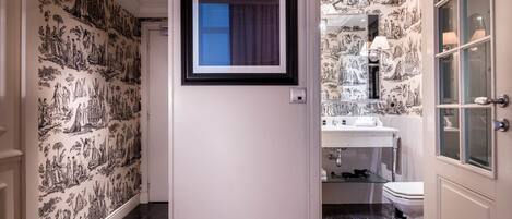 A hallway with black and white patterned wallpaper, a framed picture on the wall, and a bathroom with a sink, mirror, and toilet visible through an open door.