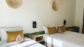 Quarto de hotel com duas camas, uma mesinha de cabeceira, uma luminária na parede e uma porta.