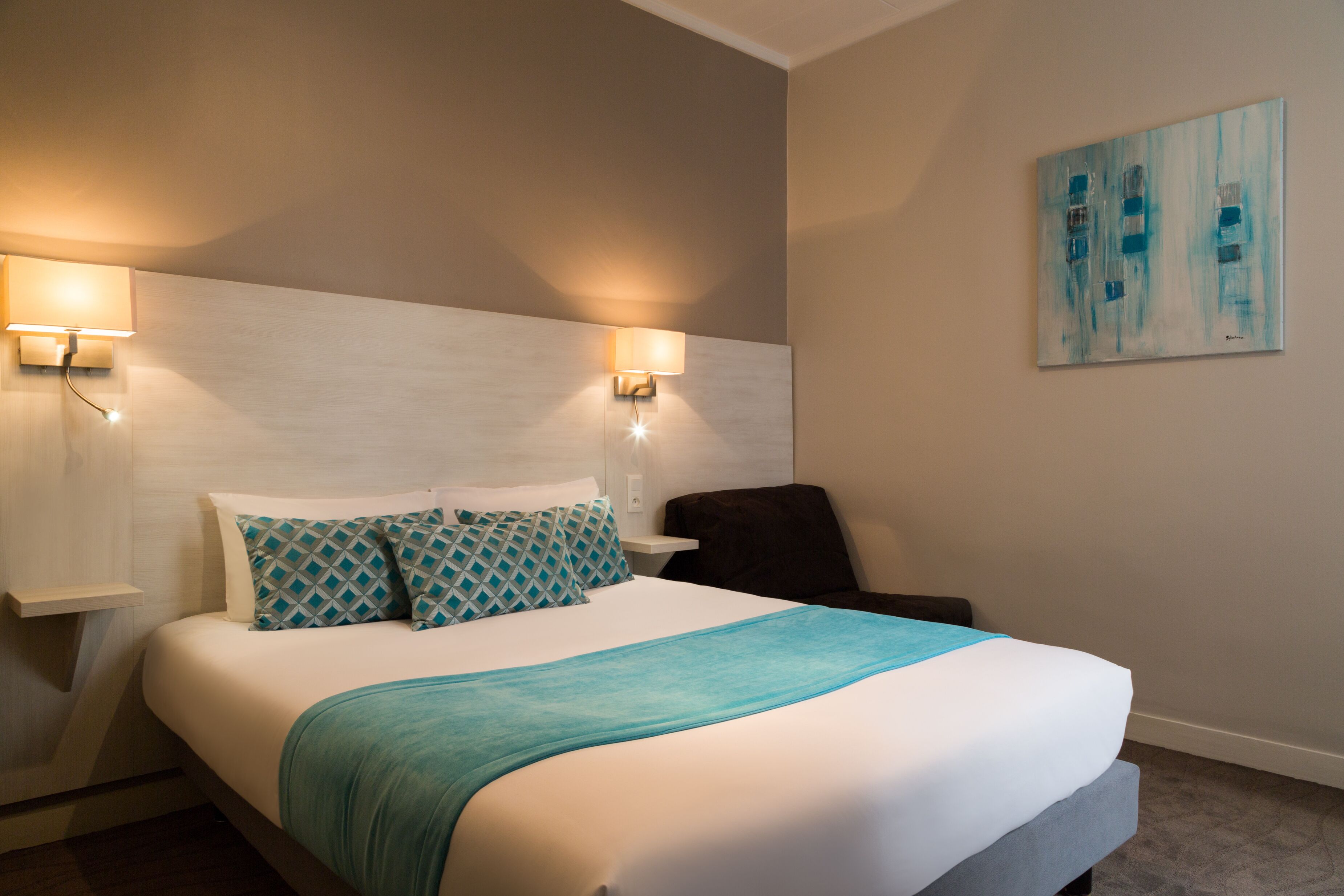 Habitación de hotel con una cama grande, dos lámparas fijadas a la pared, un sillón oscuro y un cuadro en la pared.