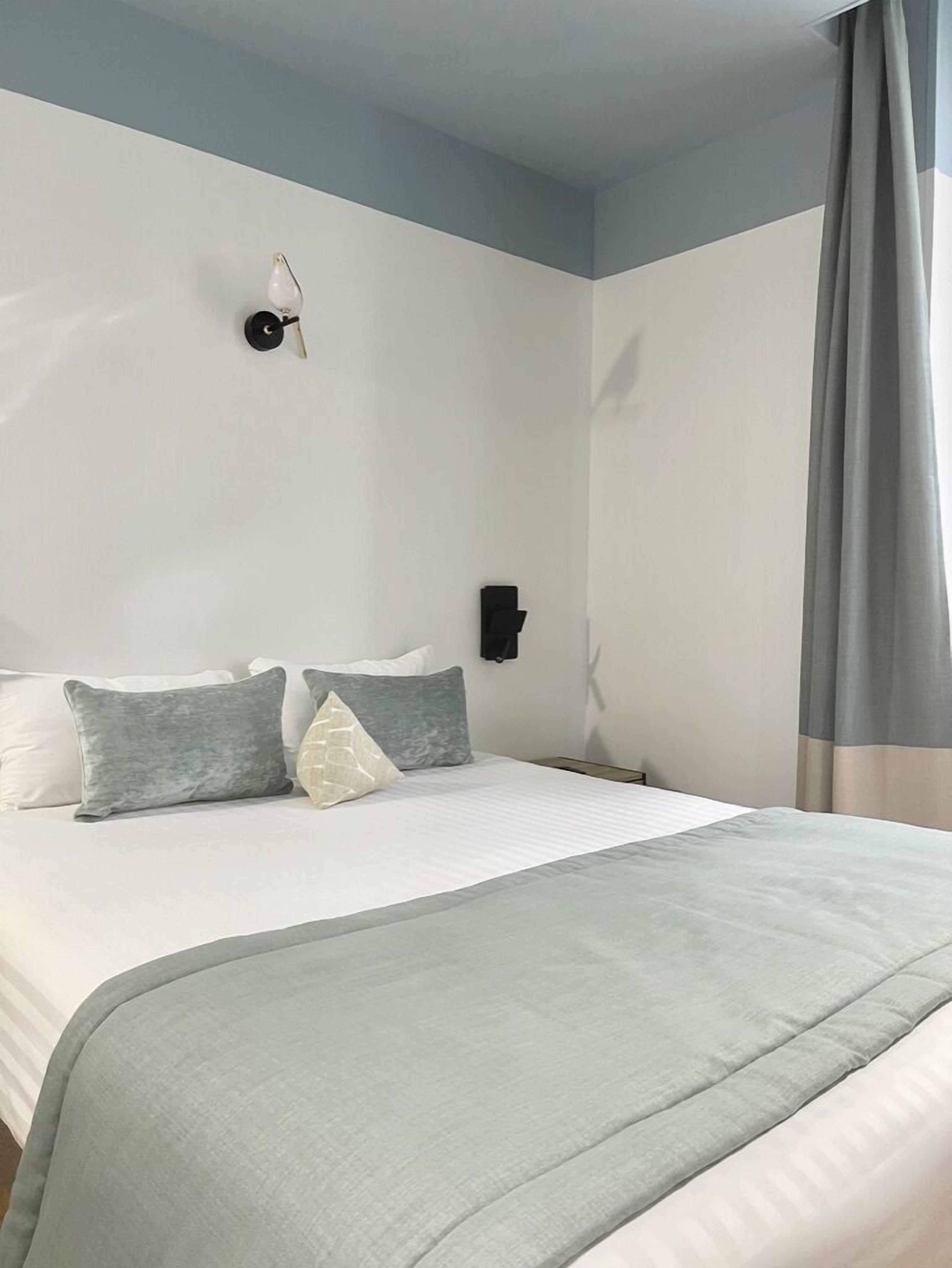 Een hotelkamer met een bed, kussens, een wandlamp en een gordijn.