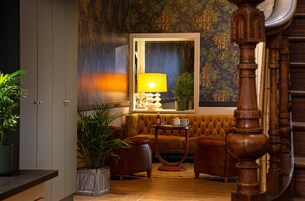 Una habitación acogedora con un sofá capitonado, una mesita redonda con una lámpara y un espejo grande que refleja un papel pintado con un motivo urbano.