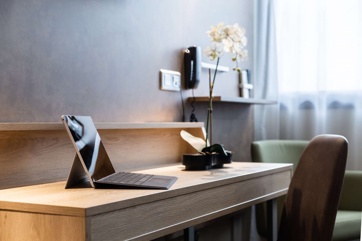 Un ordinateur portable est posé sur un bureau en bois, à côté d’un vase rempli de fleurs blanches, d’un haut-parleur noir et d’une chaise verte.