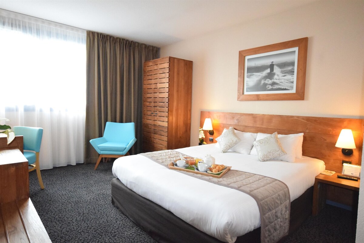 Une chambre d’hôtel avec un grand lit, une tête de lit en bois, deux lampes de chevet, un tableau encadré au mur, une chaise bleue et un bureau agrémenté d’une plante.
