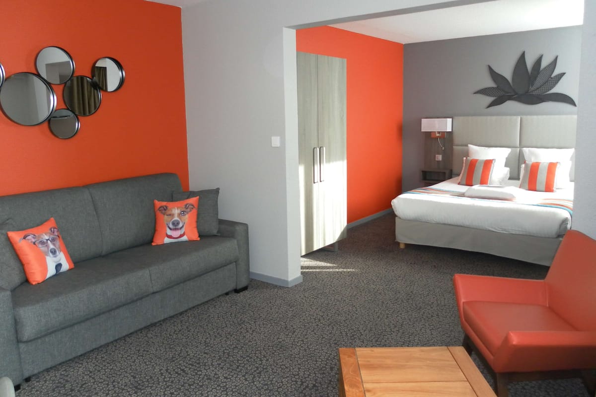 Une chambre d’hôtel avec un canapé gris, des coussins orange, une table basse en bois, un lit avec une literie blanche et orange, et un mur orné de miroirs ronds.