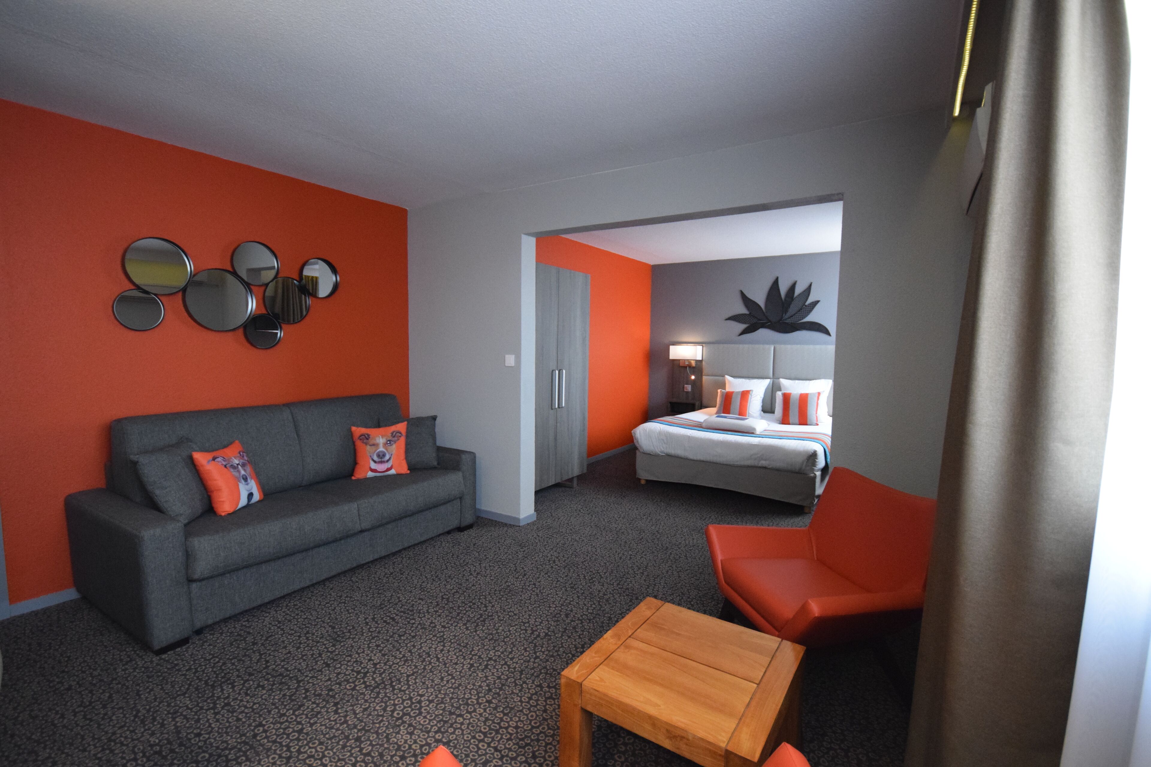 A modern hotel room with a grey sofa, orange armchair, and a wooden coffee table.
