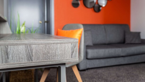 Ein modernes Wohnzimmer mit einer grauen Couch, einem Holztisch und einer leuchtend orangefarbenen Wand mit runder Wandkunst.