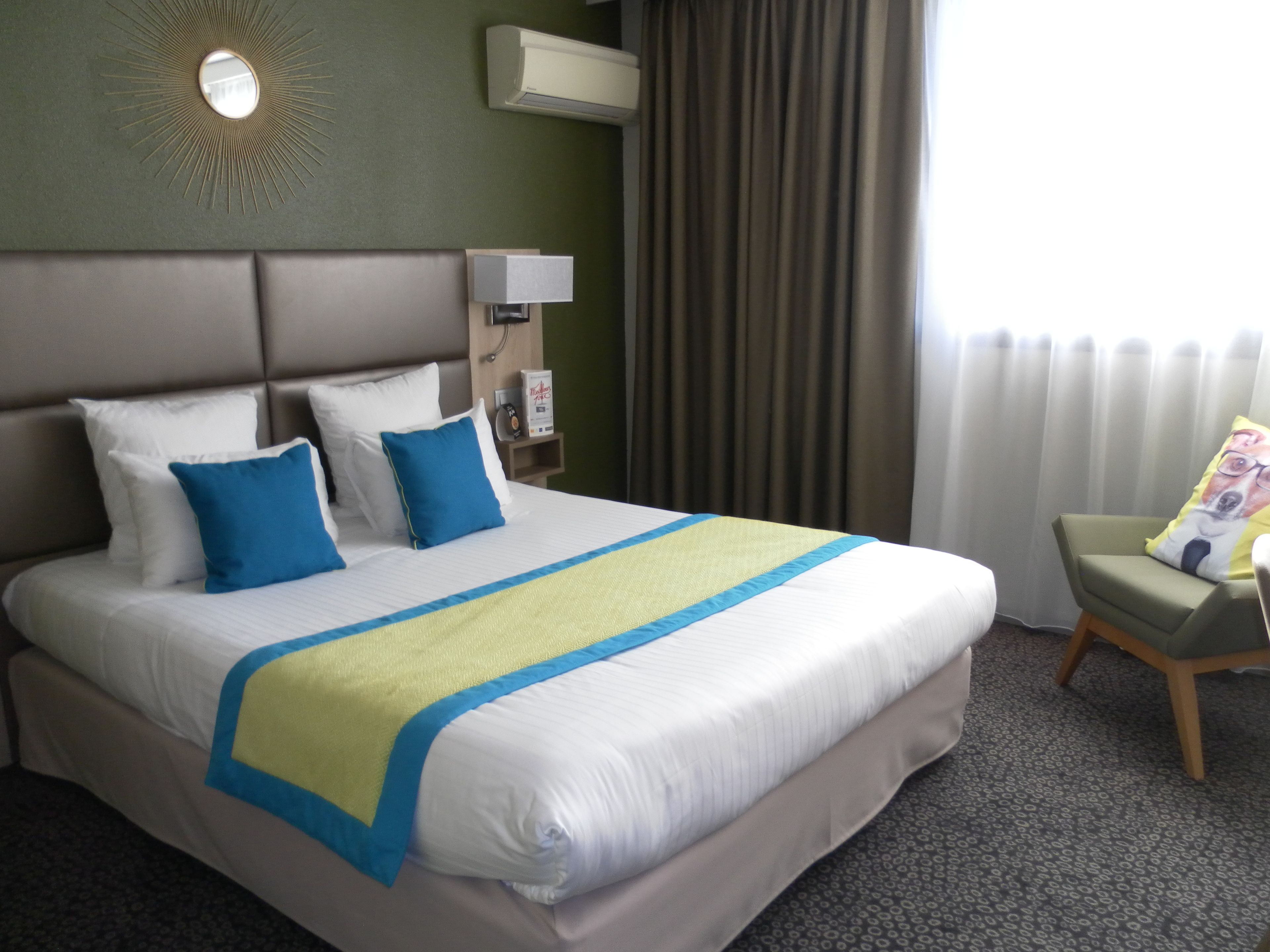 A hotel room with a large bed, a headboard, a nightstand, a lamp, a wall-mounted air conditioner, a window with curtains, and a chair with a cushion.