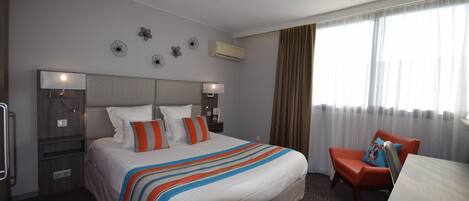 Habitación de hotel con una cama grande, dos mesitas de noche, un escritorio y una silla. Cuenta con decoraciones en las paredes y un equipo de aire acondicionado.