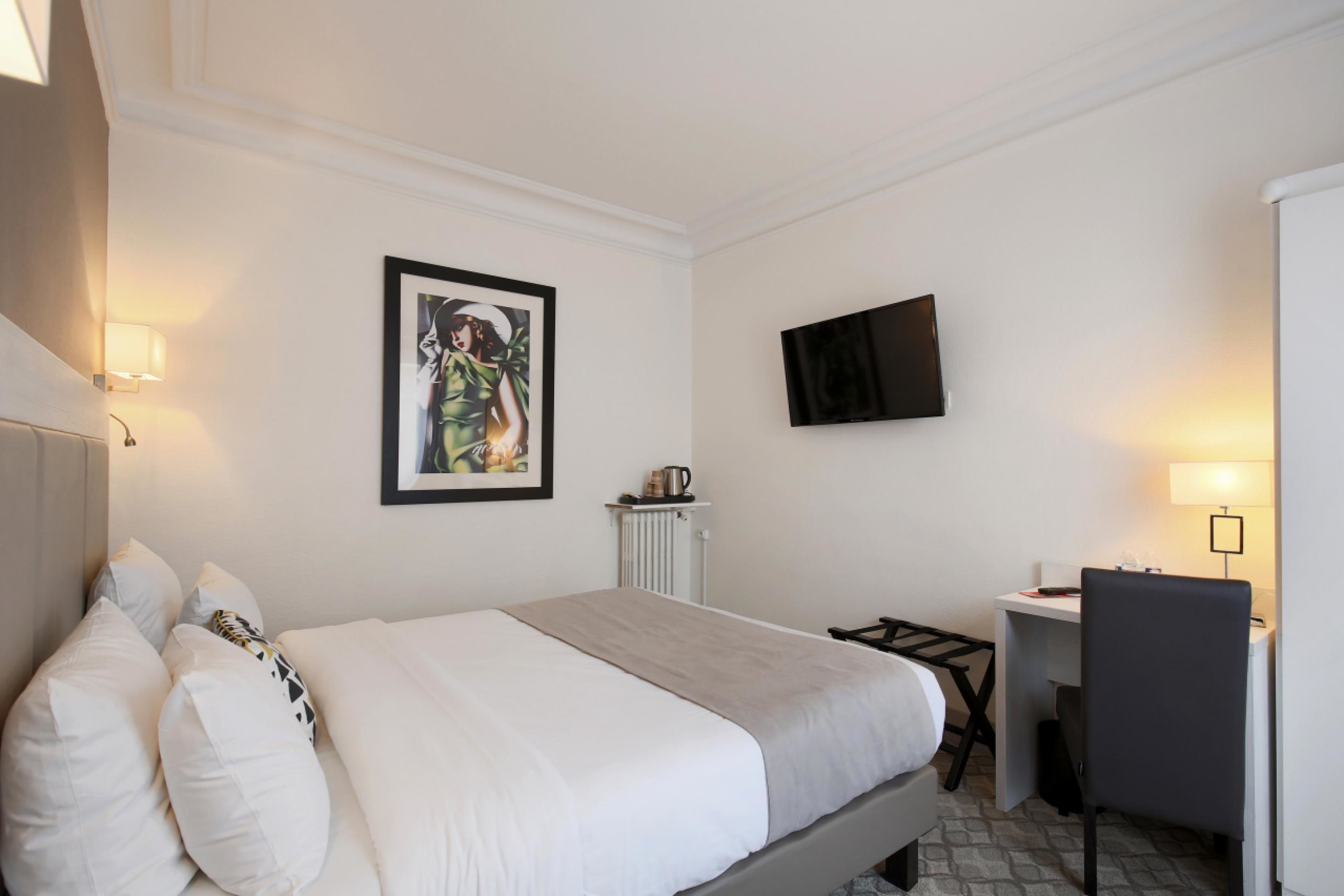 A hotel room with a bed, a desk, a chair, a TV, and a framed artwork on the wall.
