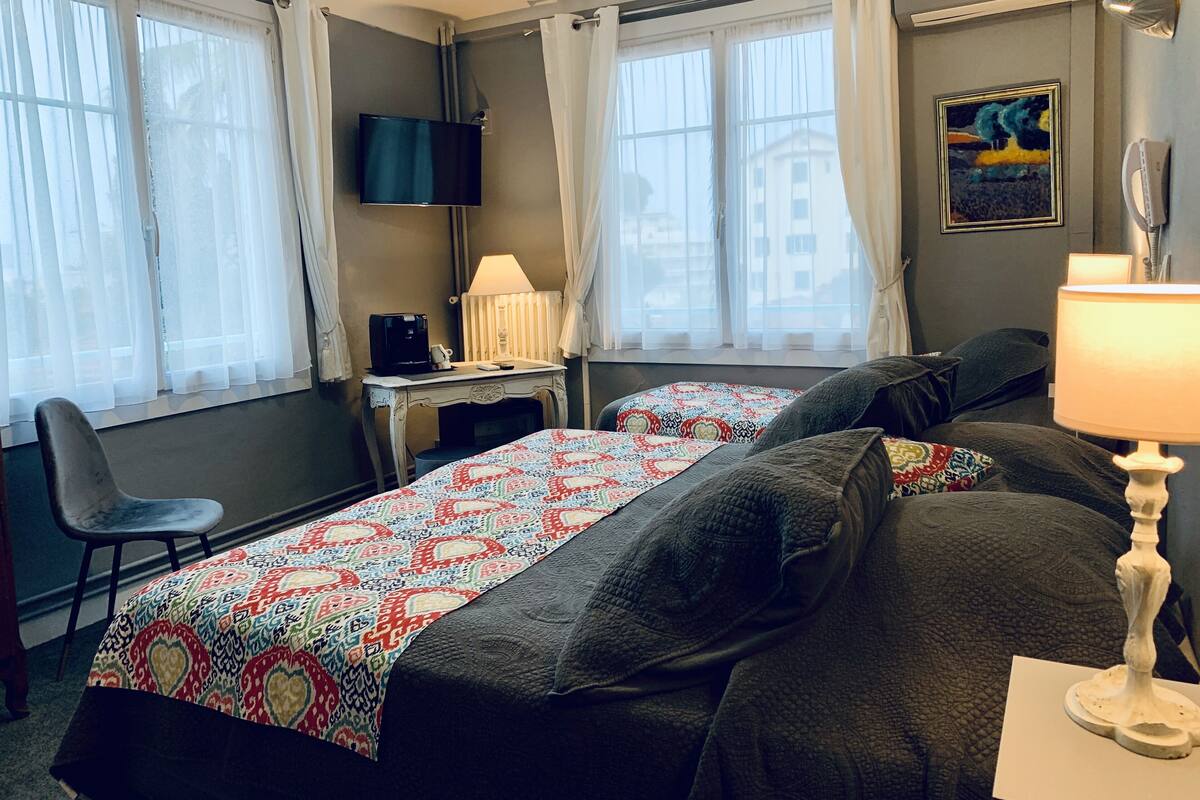 Habitación de hotel con dos camas, un escritorio con computadora y cafetera, una silla y televisor montado en la pared.