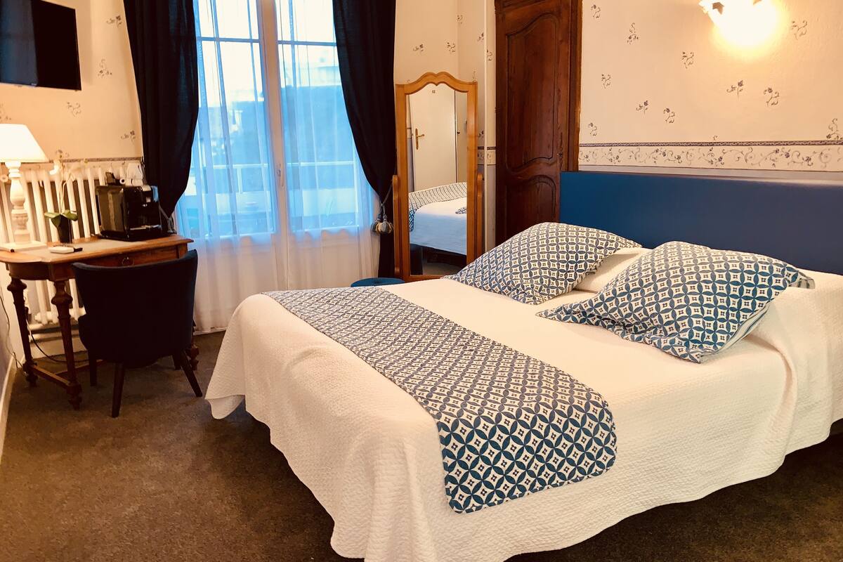 Habitación de hotel con cama, escritorio, silla, espejo y ventana con cortinas.