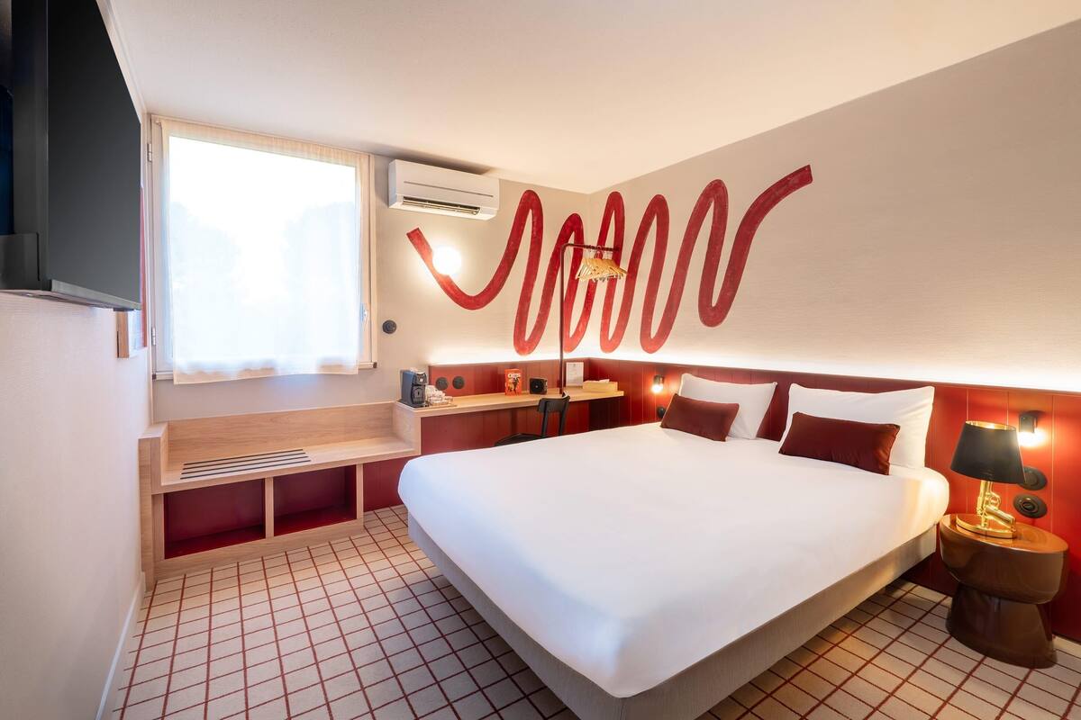 Ein Hotelzimmer mit einem großen Bett, einem Schreibtisch, einem Stuhl, einem Fenster und einer Wand-Klimaanlage.