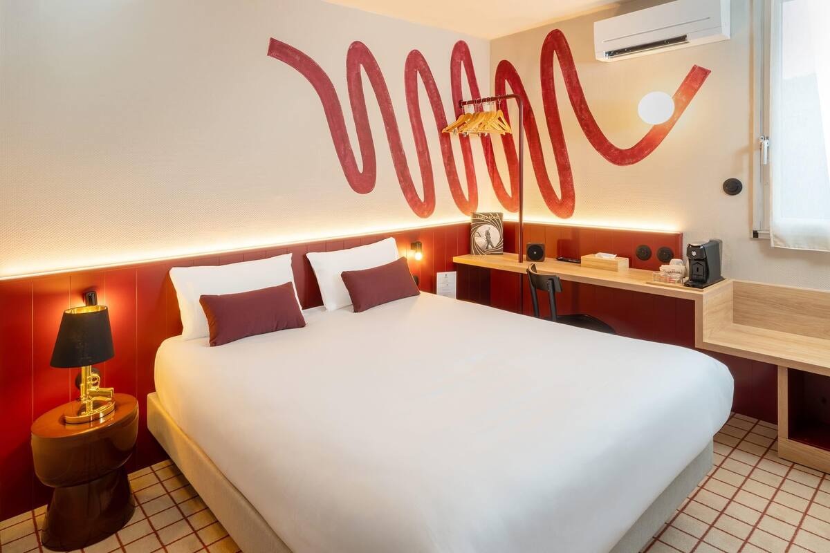 Ein Hotelzimmer mit einem großen Bett, einem Nachttisch mit Lampe, einem kleinen Schreibtisch mit Stuhl, einer Wandablage und einem großen, abstrakten Gemälde.