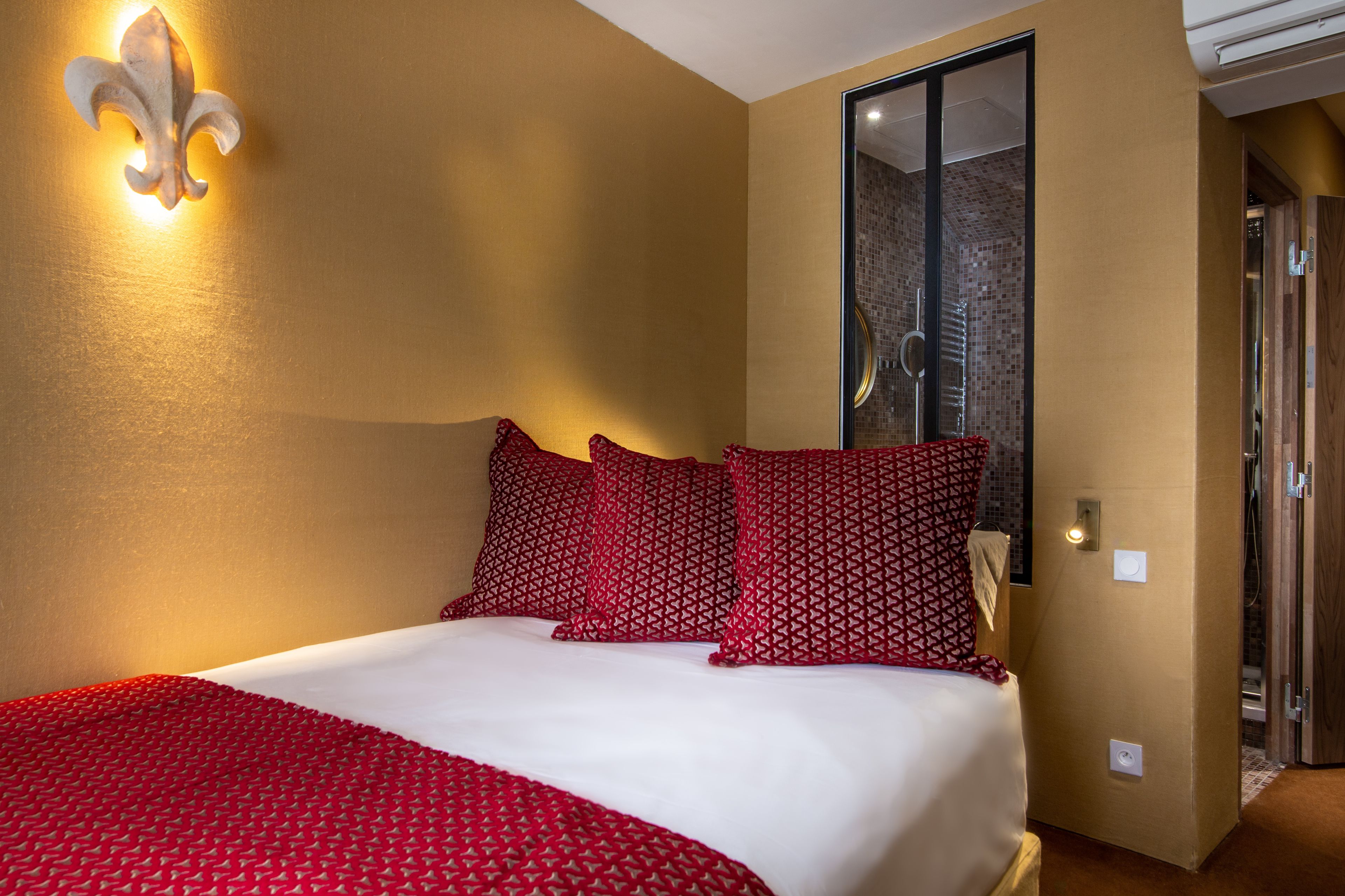 A hotel room with a bed, red patterned pillows, a wall-mounted light fixture, and a sliding glass door leading to a bathroom.