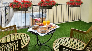 Un balcón con una mesa dispuesta para el desayuno, dos sillas de mimbre y jardineras con flores rojas.