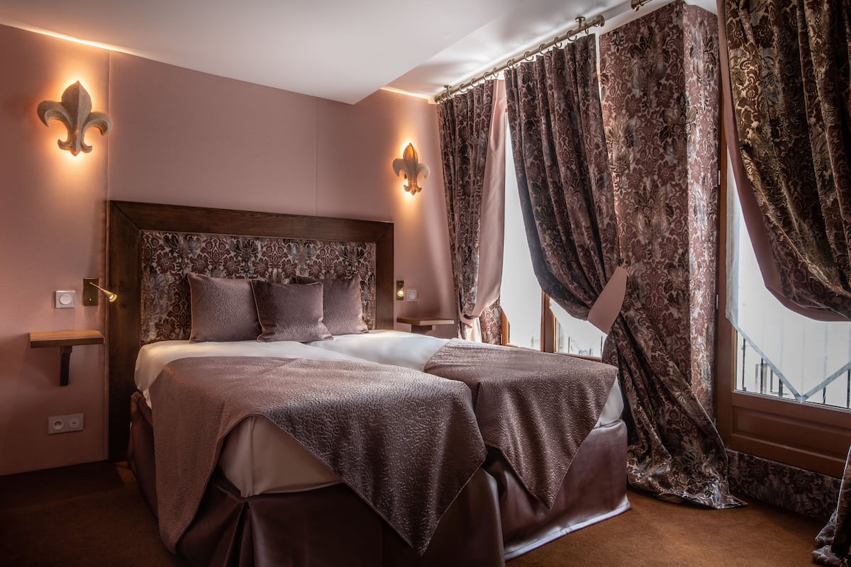 Une chambre à coucher avec un grand lit, deux tables de chevet et une tête de lit ornée d’un motif floral. Il y a des rideaux à la fenêtre et la chambre est décorée dans des tons chauds.