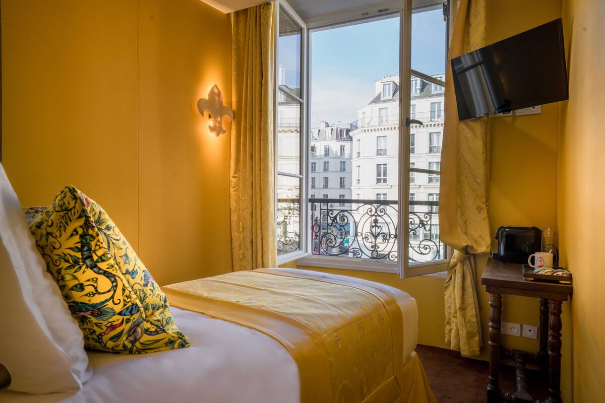 Une chambre d’hôtel avec un lit, une fenêtre donnant sur des immeubles, un téléviseur à écran plat fixé au mur et une petite table avec une machine à café.