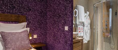 A modern hotel room with a purple textured wall, a wooden headboard, a bed with white linens, a small bedside table, and a glass-enclosed shower area with a white sink and towel rack.