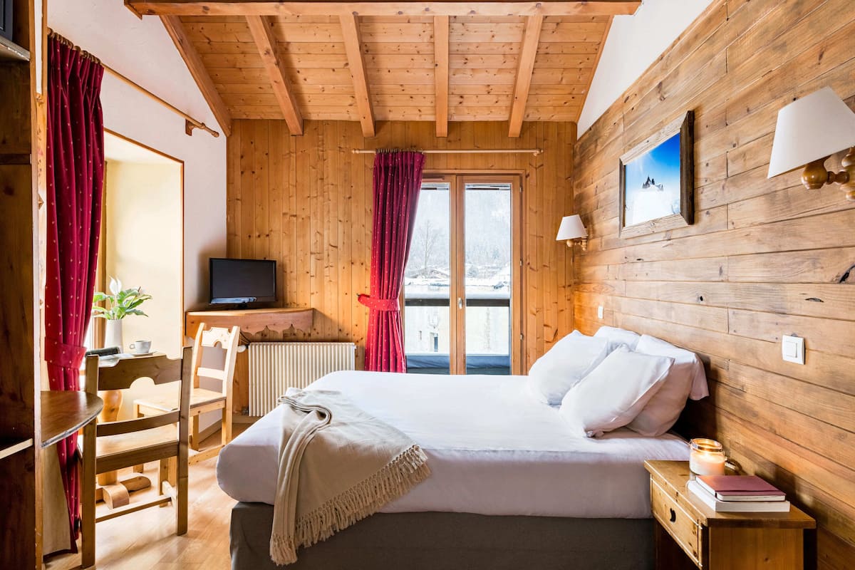 A wooden cabin room with a bed, a TV, a window with curtains, and a lamp.