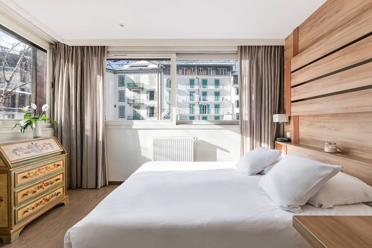 A hotel room with a large bed, wooden headboard, a dresser with a floral pattern, and a view of buildings through the window.
