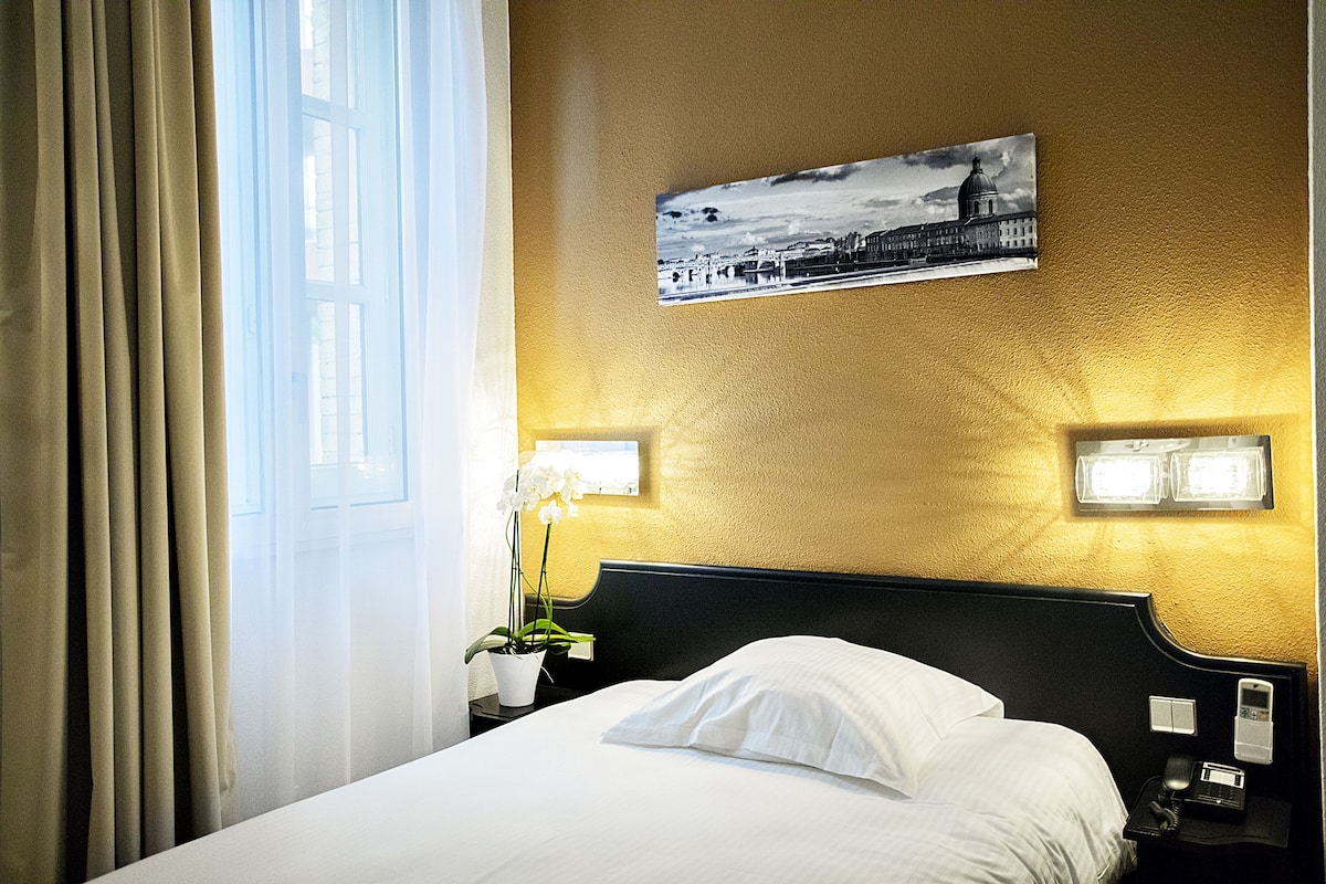 A hotel room with a bed, a nightstand with a lamp, a wall-mounted picture, and a window with curtains.