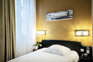 Une chambre d’hôtel avec un lit, une table de chevet avec une lampe, un tableau accroché au mur et une fenêtre avec des rideaux.