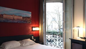 A hotel room with a bed, a painting of a building, a window with a view of a cityscape, and a nightstand with a lamp. - Occitania Centre Toulouse Matabiau (Toulouse)