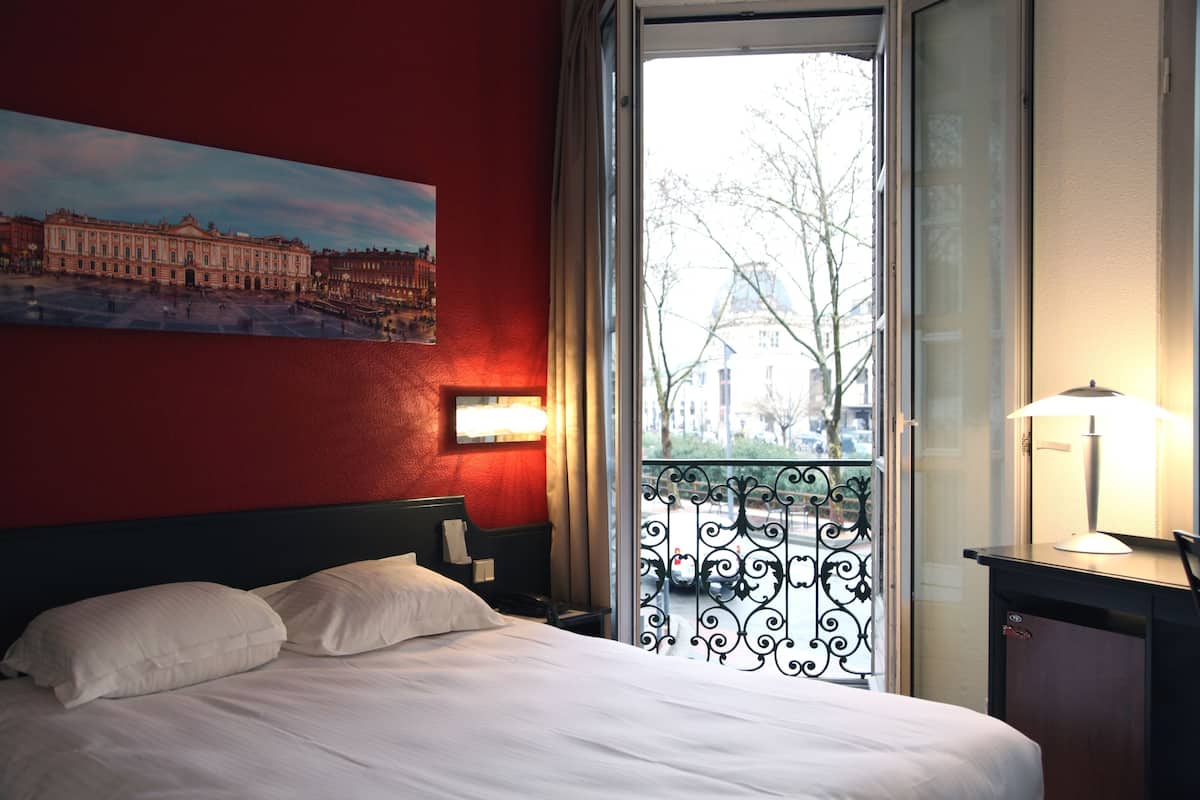 A hotel room with a bed, a painting of a building, a window with a view of a cityscape, and a nightstand with a lamp.
