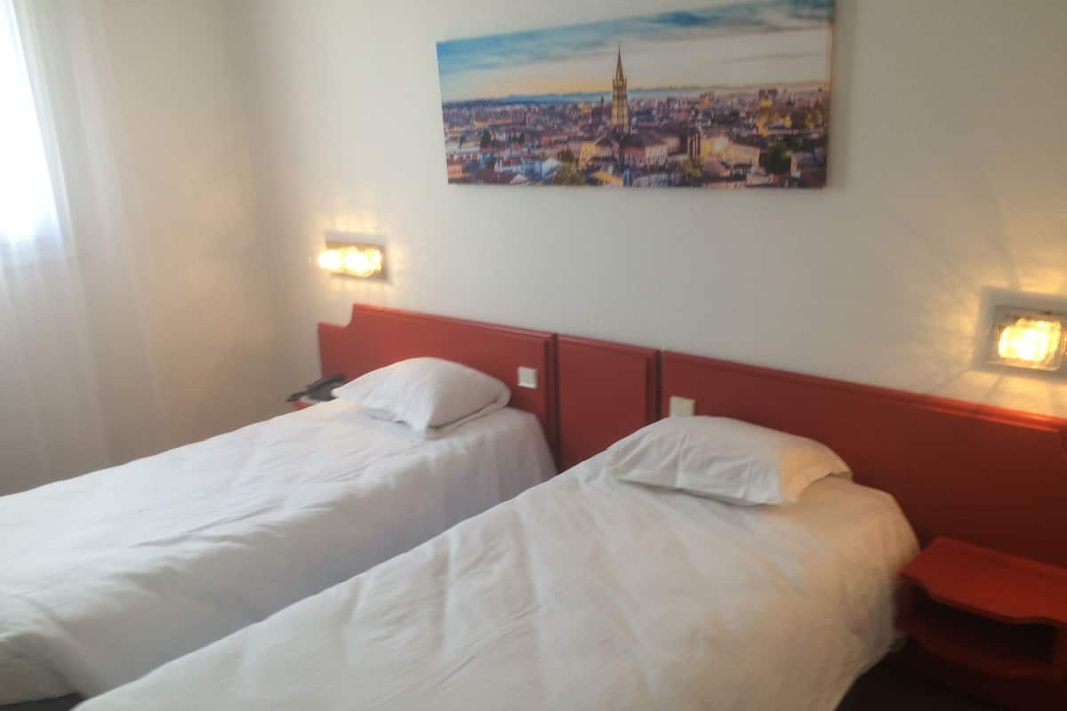 A hotel room with two single beds, a red headboard, a nightstand with a phone, and a painting of a cityscape on the wall.