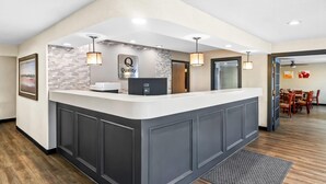 A reception area with a modern design, featuring a reception desk, a sign that reads "Quality by Choll Hotels," and a dining area visible in the background.