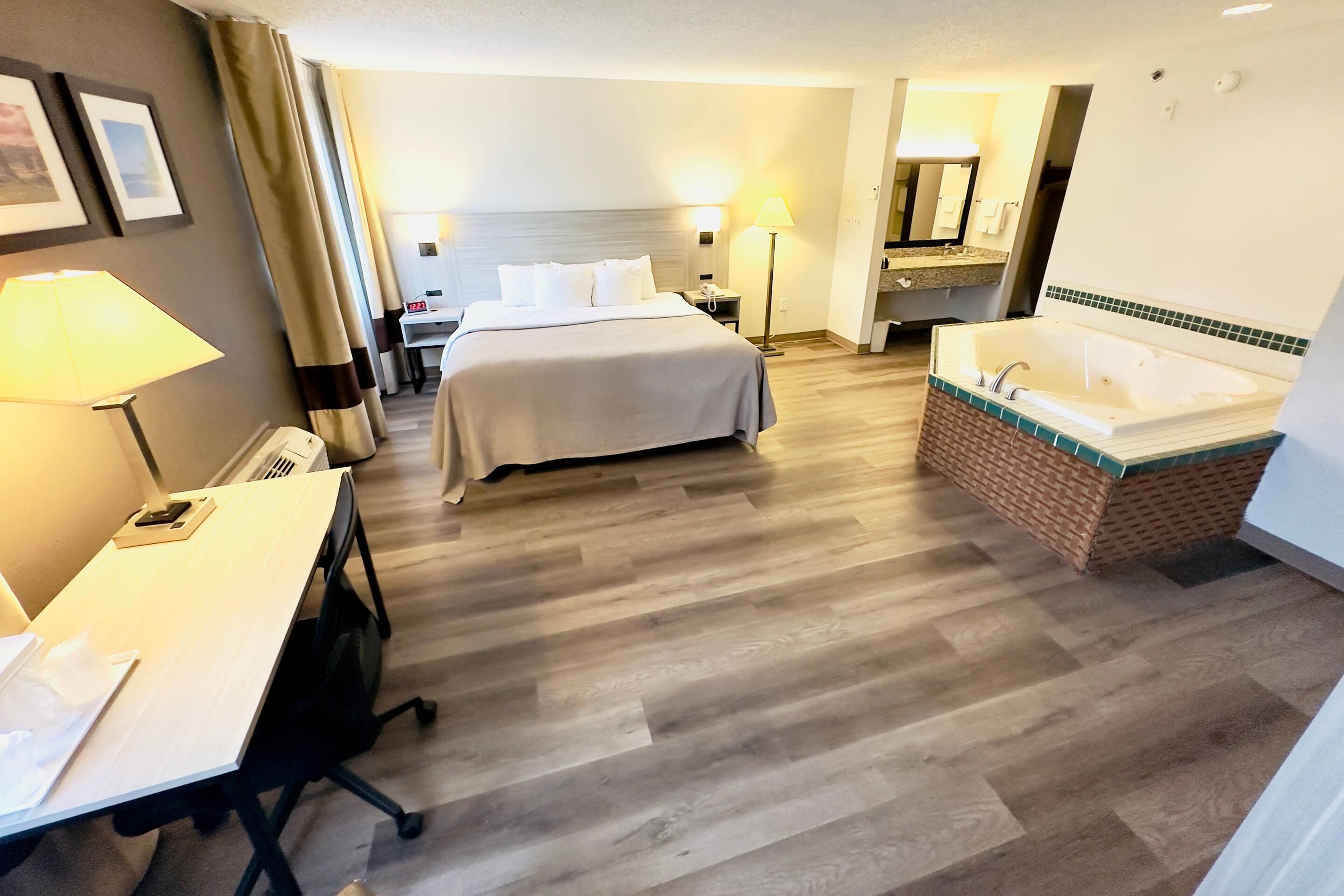 Quarto de hotel com cama, escrivaninha, luminária, banheiro com banheira e espelho.