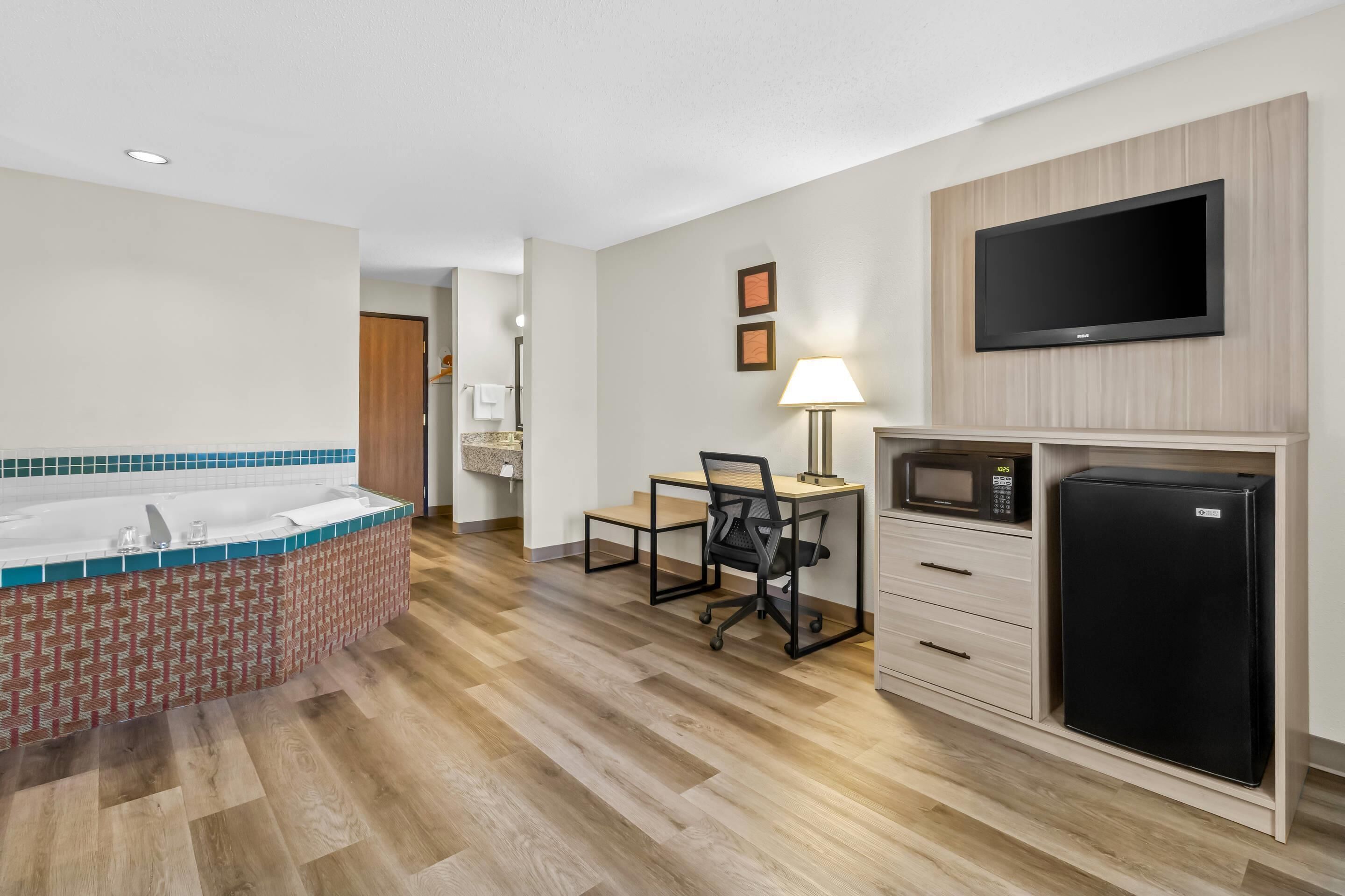 Quarto de hotel compacto com cozinha compacta, uma pequena escrivaninha com cadeira, TV de tela plana na parede, micro-ondas e geladeira.