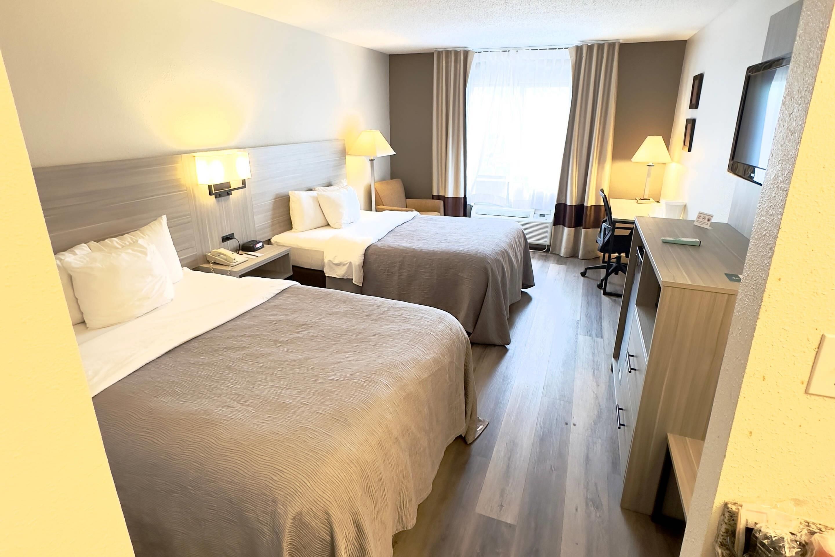Quarto de hotel com duas camas, uma mesa, uma cadeira, uma televisão e uma janela com cortinas.