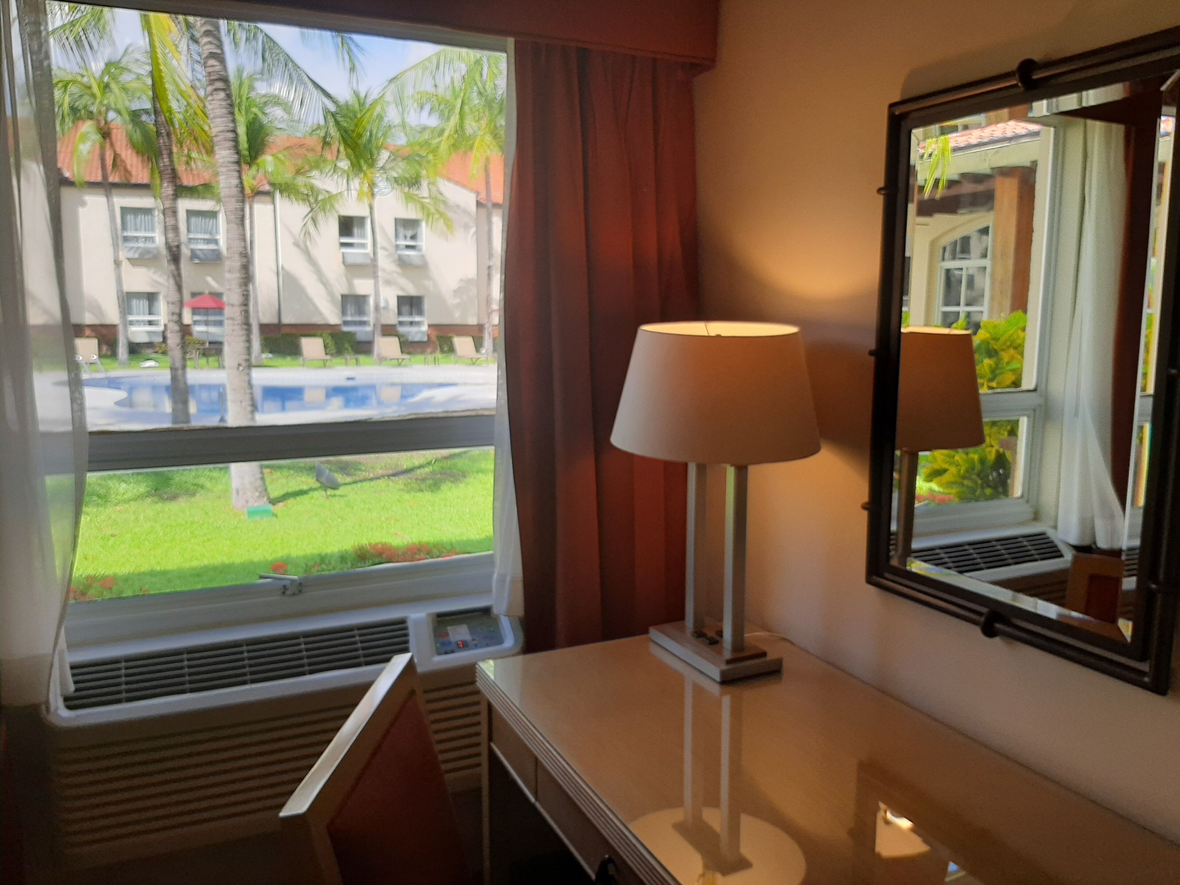 Une chambre d’hôtel avec une fenêtre donnant sur des palmiers et une piscine, une table de chevet avec une lampe et un miroir qui reflète la pièce.