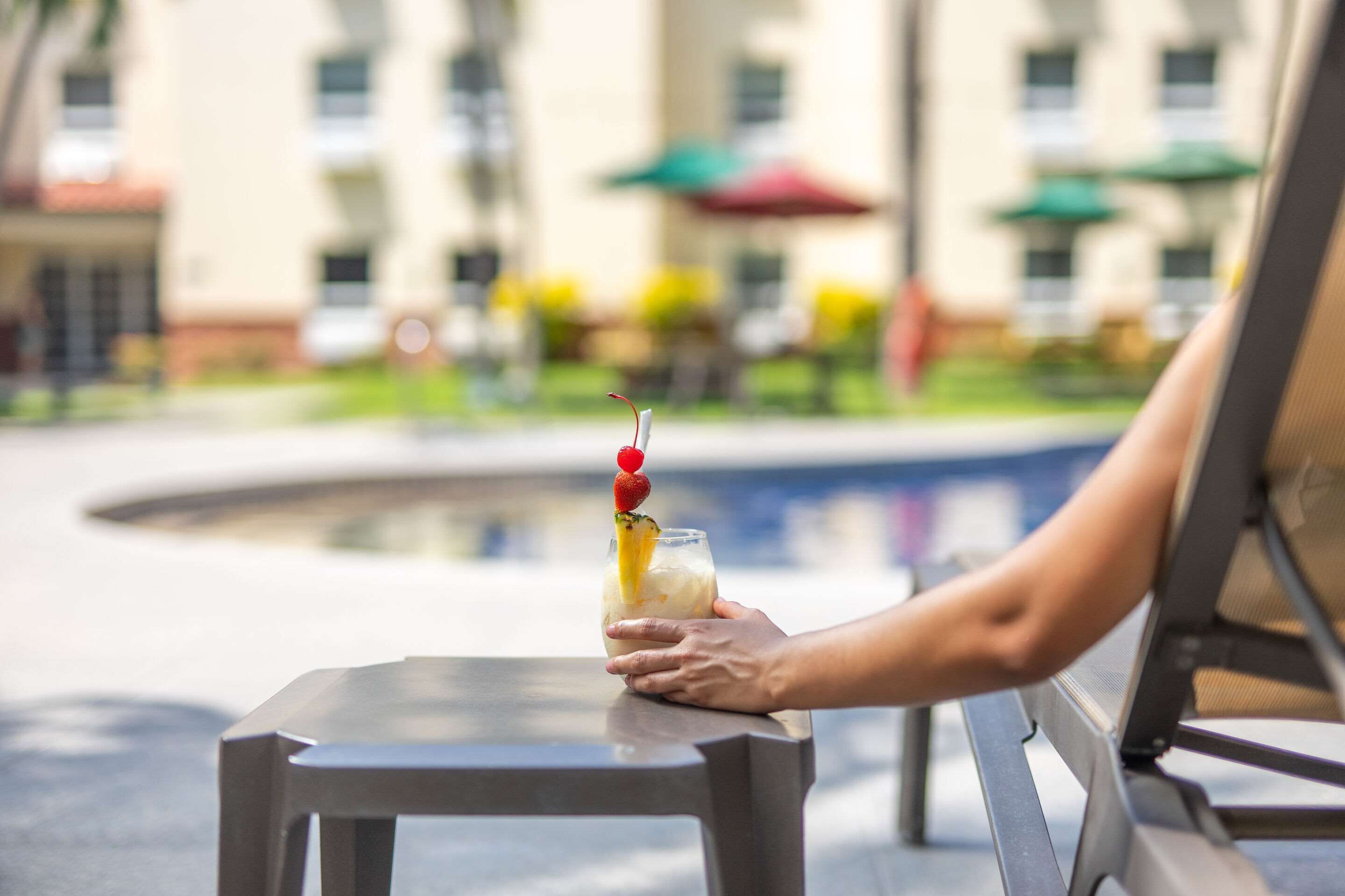 A person holding a cocktail with a cherry on a table by a poolside.