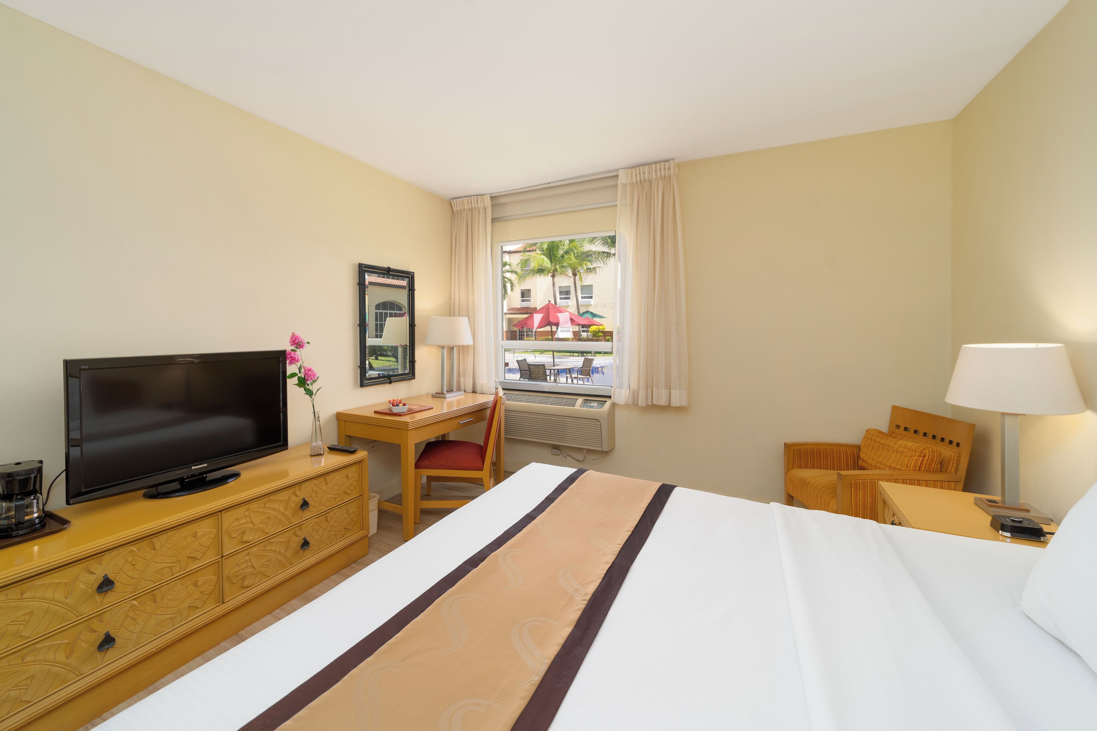 A hotel room with a bed, a TV, a desk with a chair, a window with a view of a building and palm trees, and a lamp.