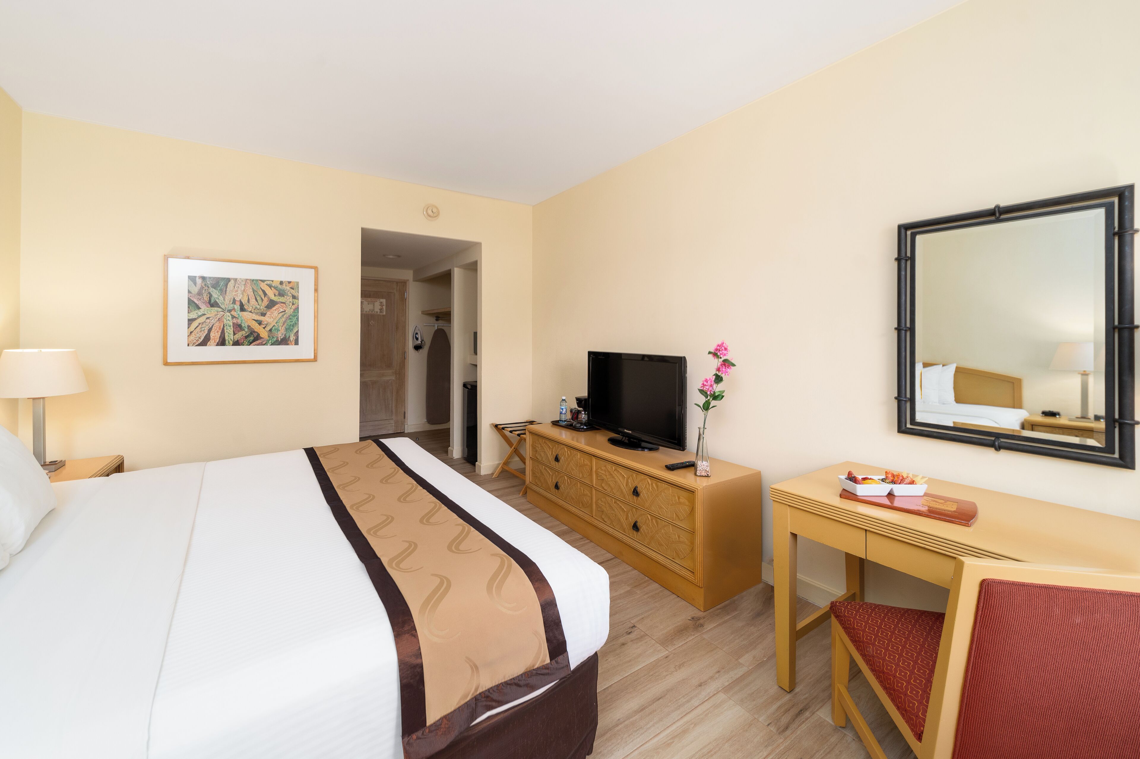 A hotel room with a bed, a TV on a wooden dresser, a desk with a chair, and a mirror on the wall.