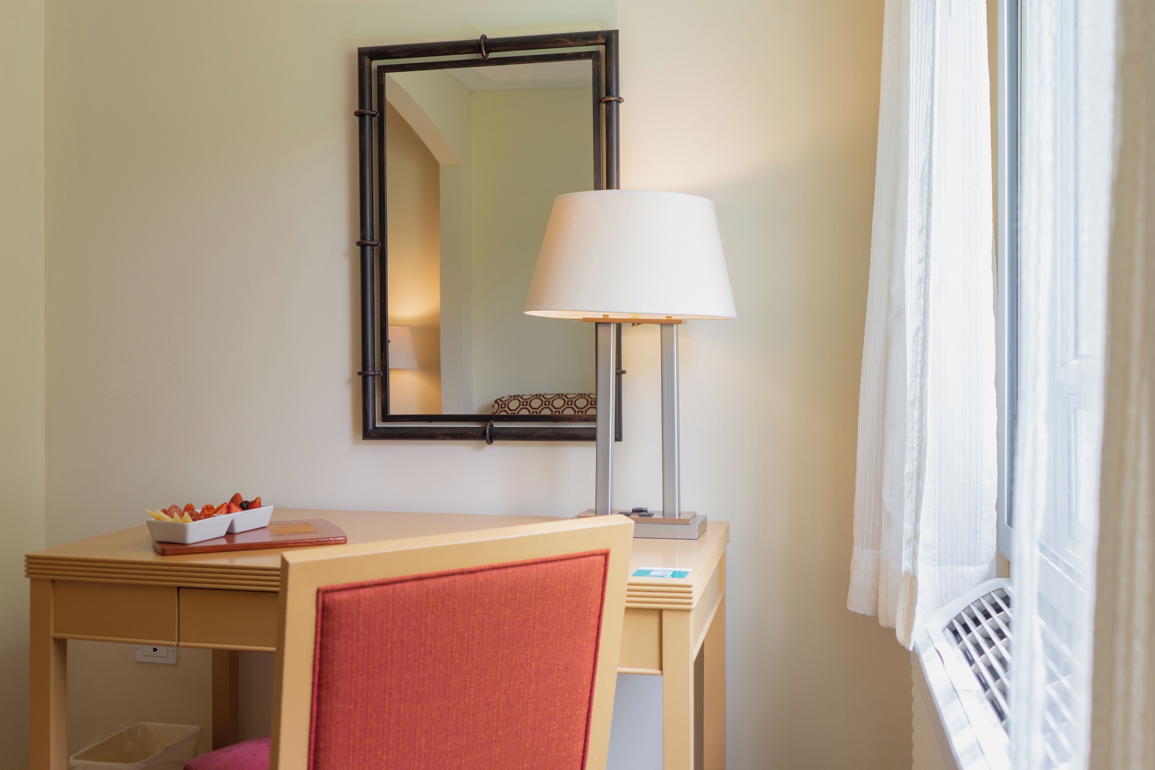Una habitación de hotel con un escritorio de madera, un espejo, una lámpara y una ventana con cortinas.