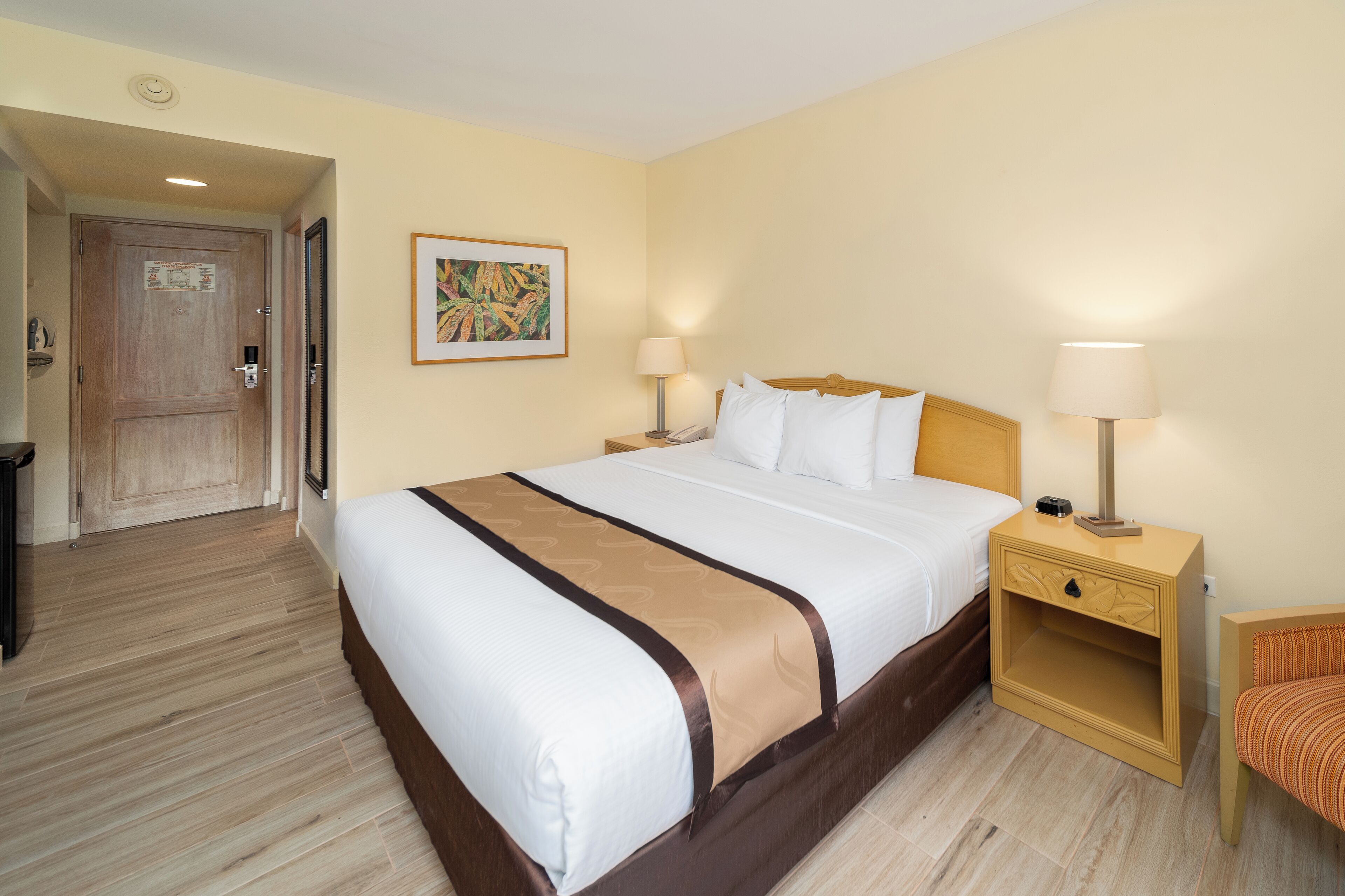 Habitación de hotel con una cama grande, dos mesitas de noche con lámparas, una silla de madera y una puerta que da a otra habitación.