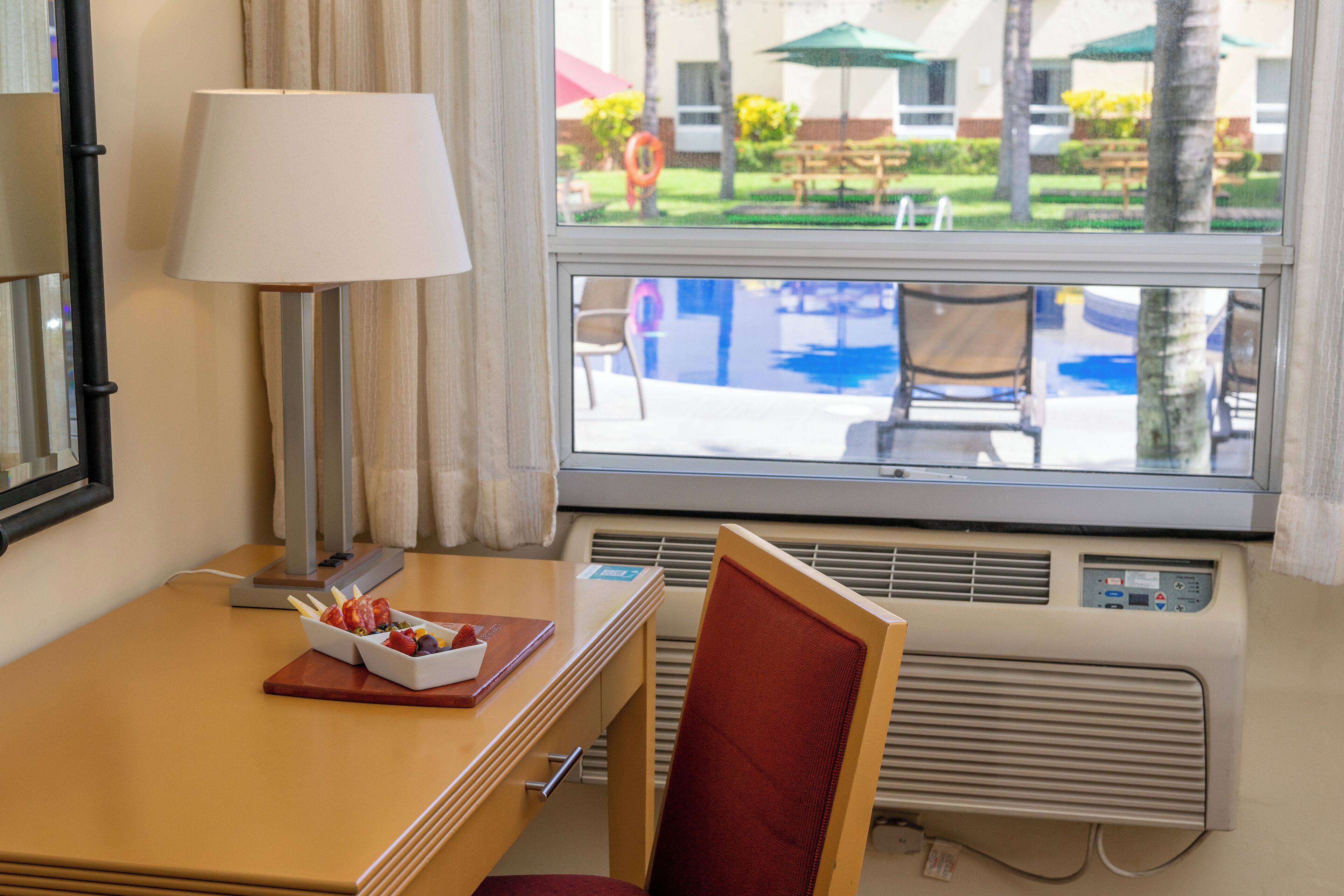 A hotel room with a wooden desk, a lamp, a chair, and a window with a view of a pool and outdoor seating.