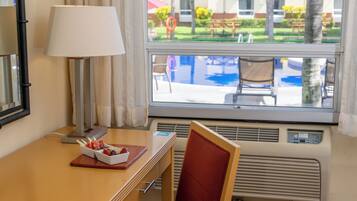 A hotel room with a wooden desk, a lamp, a chair, and a window with a view of a pool and outdoor seating.